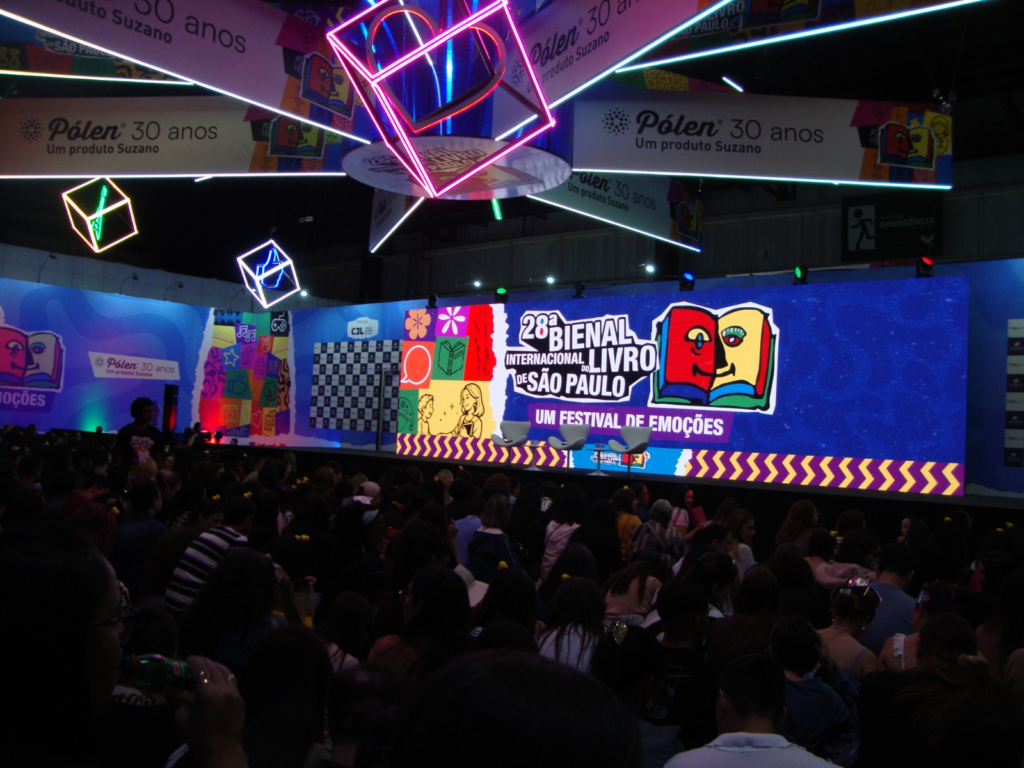 A imagem mostra a Arena Literária, um espaço que tem um grande palco com um telão de cores fortes que, no momento da foto, apresenta a logo da bienal do livro sp 2026. A platéia está localizada abaixo do palco.