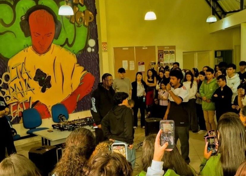 Fotografia em plano geral de uma batalha de rima em espaço comunitário iluminado por lâmpadas pendentes. Ao centro, um jovem segura o microfone falando para o público, enquanto um DJ opera uma mesa de som ao seu lado. A parede ao fundo é decorada com um grande grafite amarelo e verde. Em primeiro plano, a plateia é vista de costas, com algumas pessoas erguendo celulares para gravar a cena. é uma batalha de rima no interior de são paulo