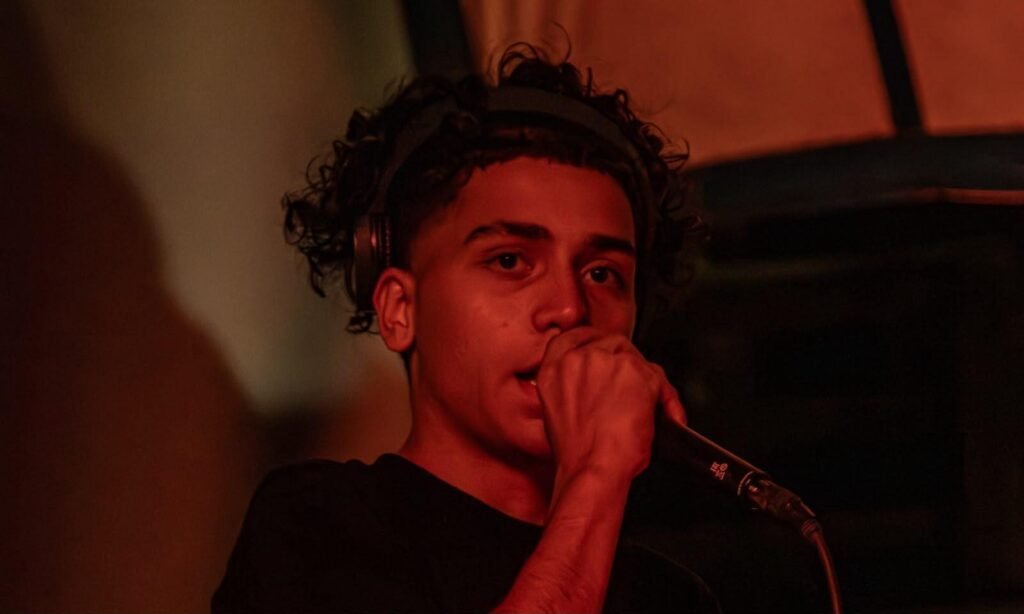 Fotografia em close-up de um jovem cantando ou se apresentando com um microfone em ambiente com iluminação vermelha e dramática. Ele possui cabelos cacheados, usa fones de ouvido pretos em volta da cabeça e segura o microfone bem próximo à boca, direcionando o olhar concentrado para a frente.