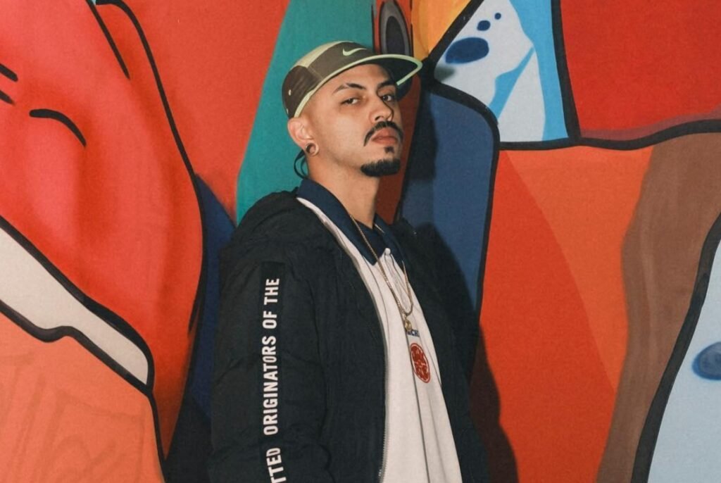 Fotografia em plano médio de um homem jovem de cavanhaque e boné bege e verde, posando no canto de uma parede decorada com um grafite colorido. Ele veste uma jaqueta preta com inscrições brancas na manga sobre uma camiseta polo branca. A parede ao fundo apresenta uma arte urbana em estilo pop art com traços pretos marcantes e blocos de cores vivas como vermelho, laranja e azul.