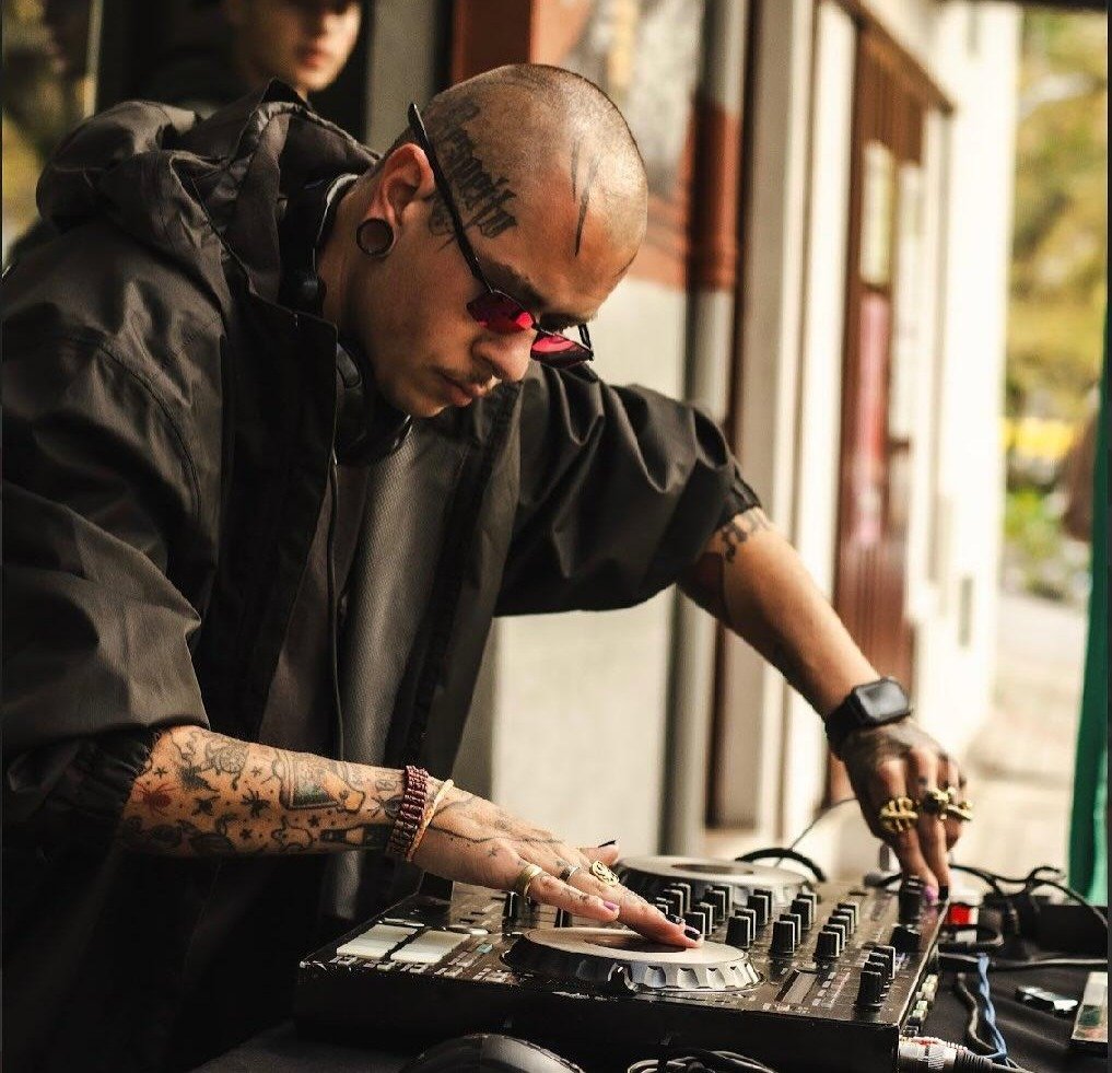 Fotografia em plano médio de um DJ de cabeça raspada e tatuagens no rosto e nos braços, operando uma controladora de áudio em um evento ao ar livre. Ele usa óculos escuros com armação pequena, fones de ouvido ao redor do pescoço e uma jaqueta escura. Seus dedos estão posicionados sobre os botões do equipamento de som. Ao fundo, desfocado, outro rapaz de boné preto observa a performance.