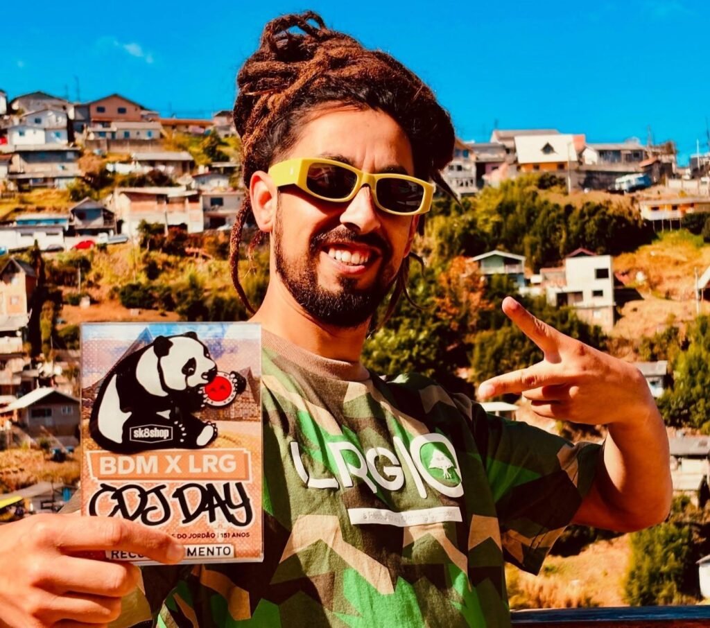 Fotografia em plano médio de um homem sorridente de dreadlocks presos e óculos de sol amarelos, posando ao ar livre sob luz solar direta. Ele veste uma camiseta camuflada verde e faz o sinal de "paz e amor" com uma das mãos, enquanto segura com a outra um encarte impresso com a ilustração de um panda e o texto "BDM X LRG". Ao fundo, observa-se uma paisagem urbana com casas em uma região periféria em uma encosta.