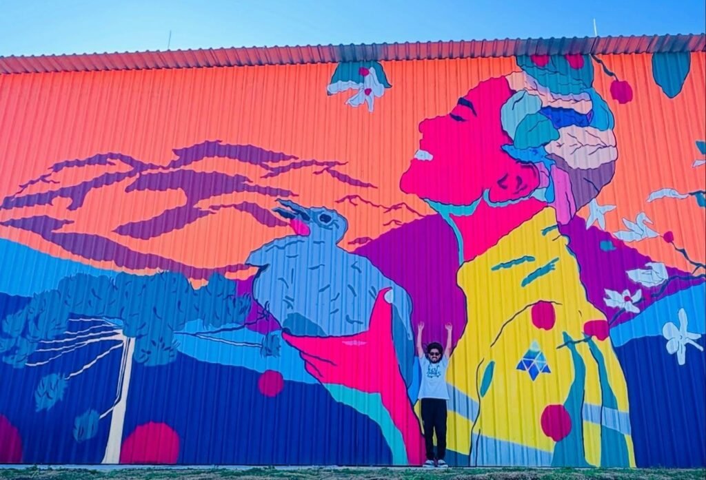 Fotografia em plano geral e ângulo baixo mostrando uma pessoa em pé com os braços erguidos em frente a um enorme mural colorido pintado na parede de uma estrutura metálica. A pintura mural traz ilustrações estilizadas de um perfil humano, pássaros, montanhas e vegetação, utilizando cores contrastantes como rosa, amarelo, azul e laranja sob um céu azul limpo. grafite localizado no interior de são paulo