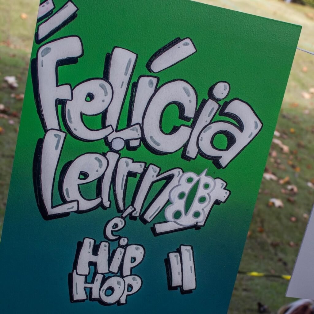 Fotografia em close-up de um cartaz verde exibido na vertical. O cartaz traz a frase "Felícia Leirner é HIP HOP" desenhada em estilo grafite, com letras gordinhas na cor cinza e contorno preto, destacando-se sobre o fundo verde com um gramado desfocado ao fundo.
