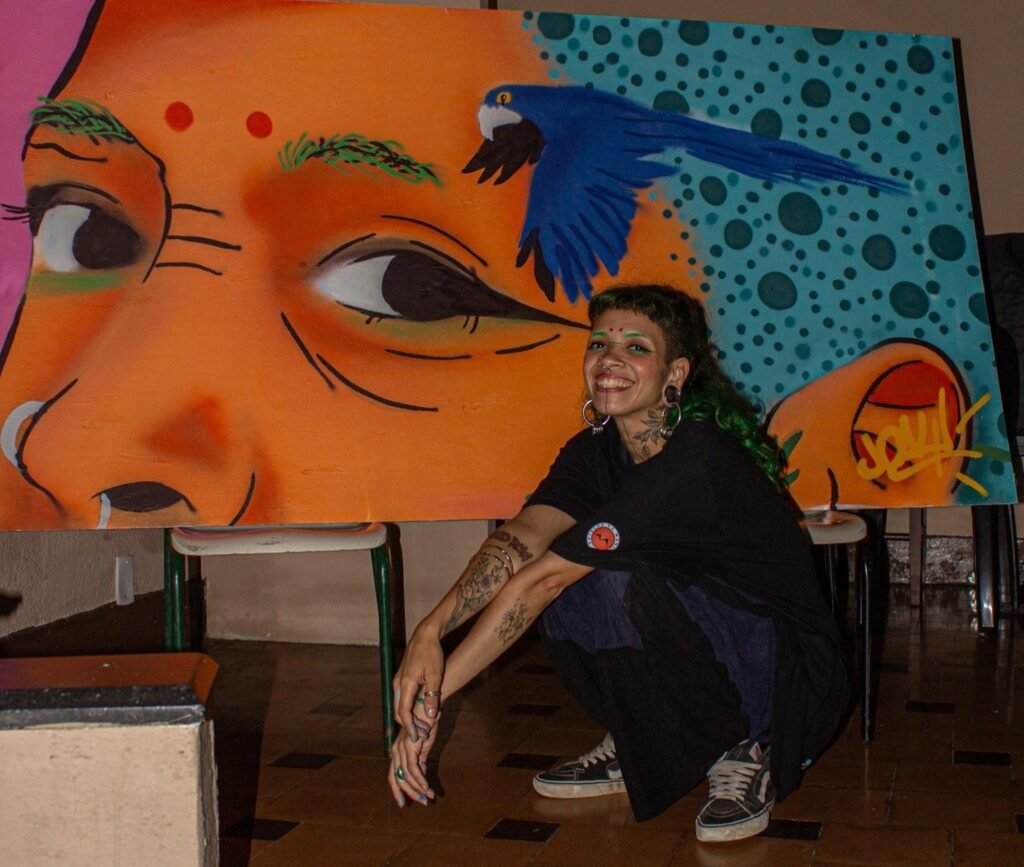 Fotografia em plano médio de uma artista agachada sorrindo ao lado de sua pintura em tela de grande formato. Ela possui cabelos escuros com franja curta e detalhes verdes, usa alargadores nas orelhas, piercings no rosto e traja roupas escuras com tênis. A pintura atrás dela exibe o rosto estilizado de uma pessoa em tons alaranjados, um pássaro azul voando ao centro e um fundo verde com bolinhas escuras. as roupas tem estilo próxicmo ao hiphop