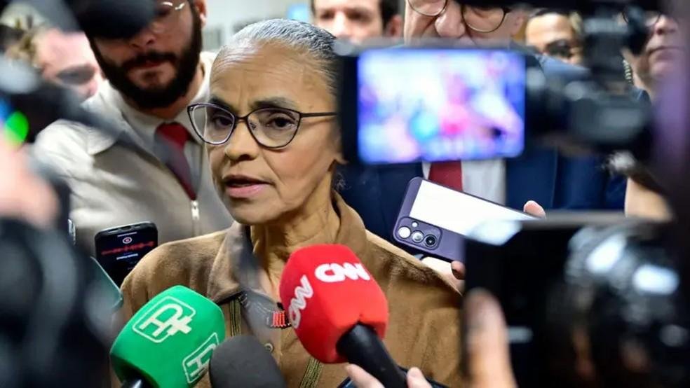 Mulher negra de coque e óculos falando com microfones