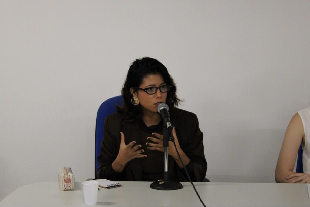 Mulher negra de óculos, blazer marrom falando ao microfone