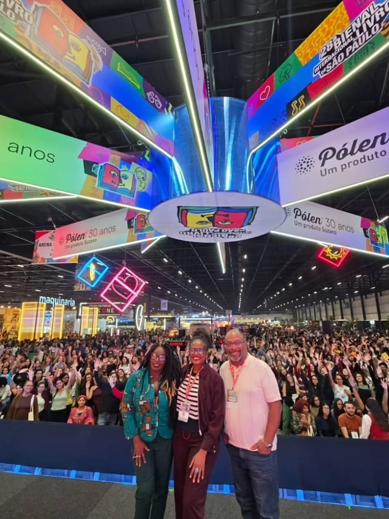 Fotografia em plano geral os palestrantes e a mediadora posando sorridentes em primeiro plano, enquanto atrás deles uma multidão de visitantes ergue os braços em sinal de celebração. O espaço amplo é decorado com grandes banners suspensos no teto referentes à "28ª Bienal Internacional do Livro de São Paulo" e patrocínios do evento.