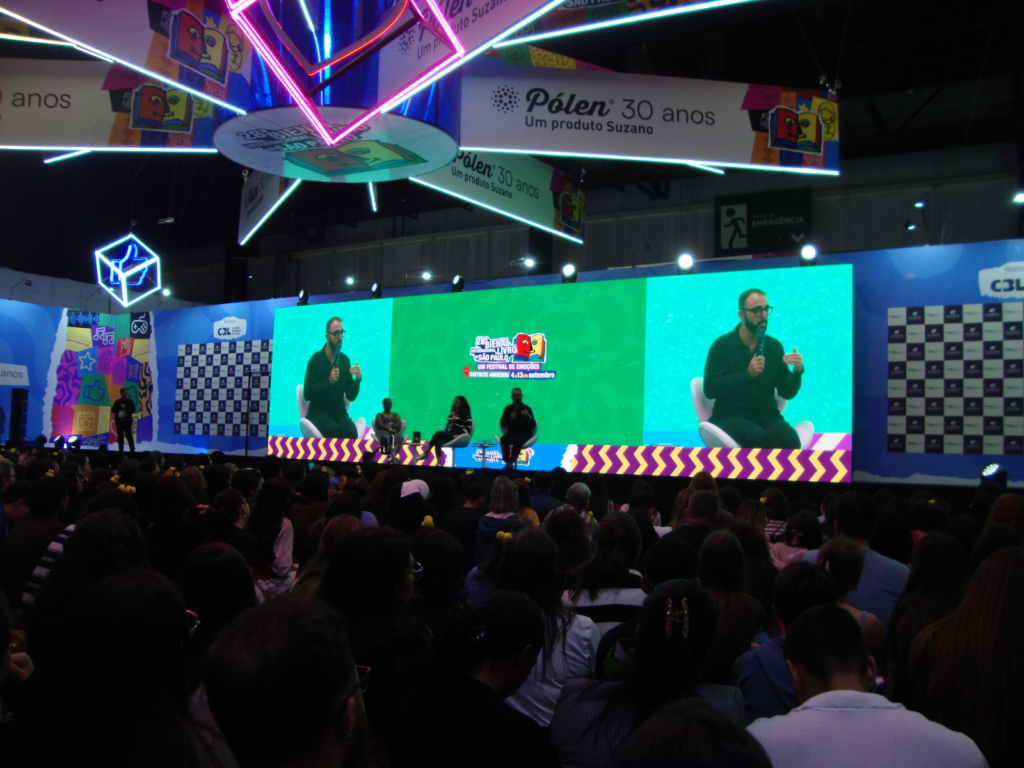 Fotografia em plano geral da plateia lotada de um auditório em uma feira literária. Ao fundo, no palco, sobressai-se um enorme telão de LED verde que exibe a transmissão ao vivo de um palestrante de óculos e camisa escura conversando com convidados no palco. Acima da estrutura do palco, banners suspensos destacam a marca "Pólen 30 anos" e a comunicação visual do evento com luzes de neon.