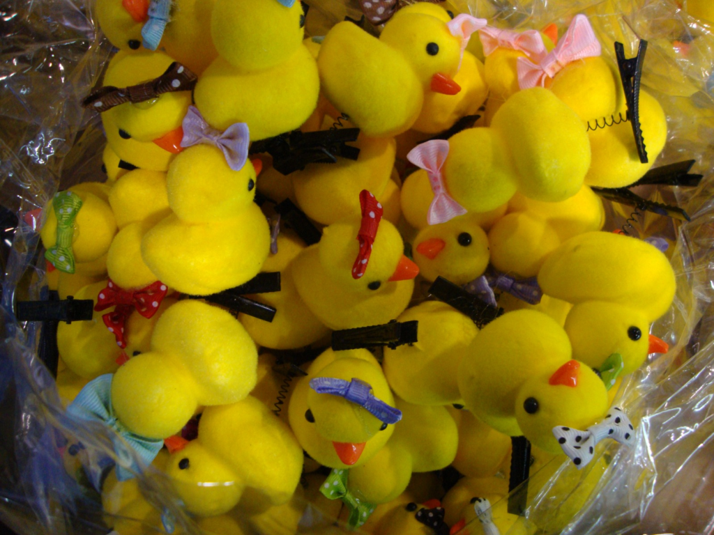 Fotografia em plano detalhe e vista superior mostrando uma pilha densa de pequenos pintinhos de pelúcia amarelos dentro de um saco plástico transparente. Cada pintinho possui um bico alaranjado e pequenos laços coloridos — em tons de rosa, roxo, vermelho com bolinhas ou azul — presos ao topo de suas cabeças por prendedores de cabelo pretos.