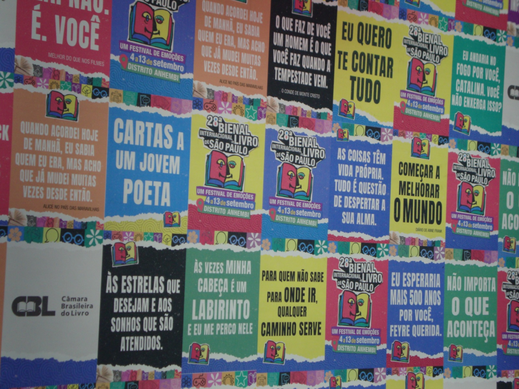 Fotografia em plano médio de uma parede inteiramente coberta por uma colagem de cartazes e painéis gráficos coloridos. Os cartazes exibem frases inspiradoras e literárias em letras garrafais brancas sobre fundos de cores variadas como verde, azul, vermelho e amarelo, intercalados com o logotipo oficial da "28ª Bienal Internacional do Livro de São Paulo".