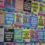 Fotografia em plano médio de uma parede inteiramente coberta por uma colagem de cartazes e painéis gráficos coloridos. Os cartazes exibem frases inspiradoras e literárias em letras garrafais brancas sobre fundos de cores variadas como verde, azul, vermelho e amarelo, intercalados com o logotipo oficial da "28ª Bienal Internacional do Livro de São Paulo".