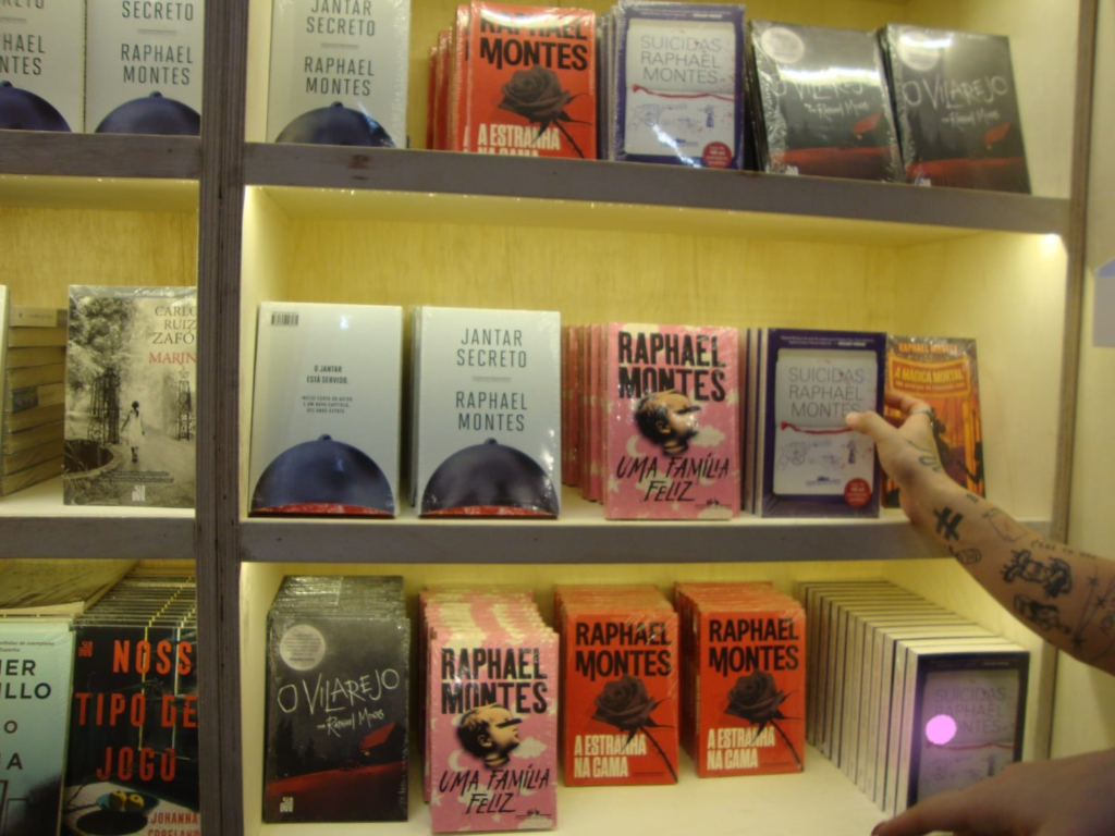 Fotografia em plano detalhe de uma estante de livraria iluminada por fitas de LED amareladas, repleta de exemplares de romances do autor Raphael Montes. Na prateleira do meio, uma mão com tatuagens nos dedos e no braço alcança a capa do livro "Suicidas", posicionado ao lado de outros títulos conhecidos como "Jantar Secreto" e "Uma Família Feliz".