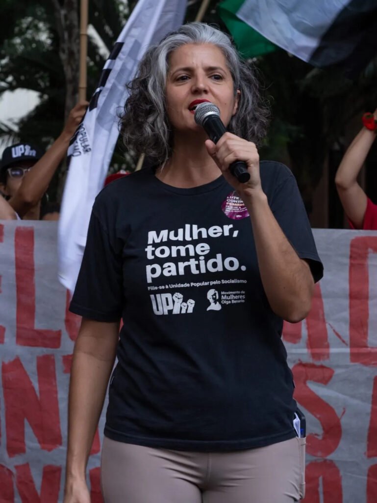 Mulher branca de cabelo branco e camiseta preta falando no microfone