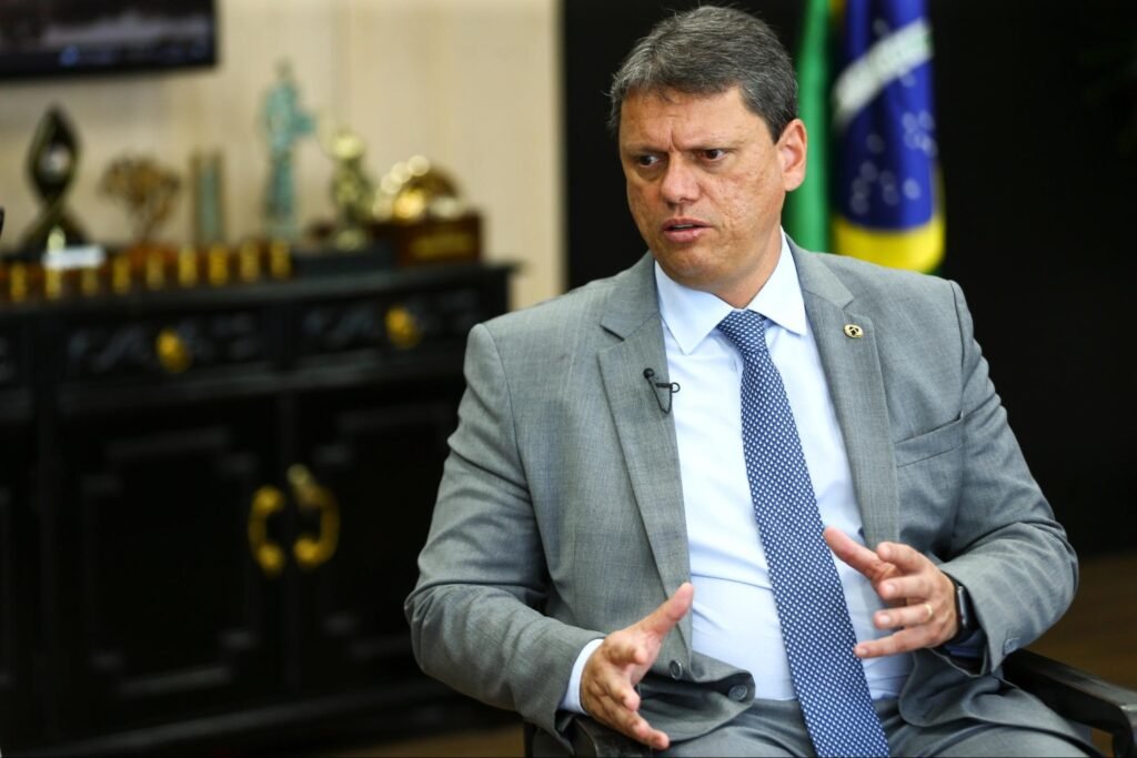 Homem branco de cabelo acizentado com terno cinza e gravata azul sentado