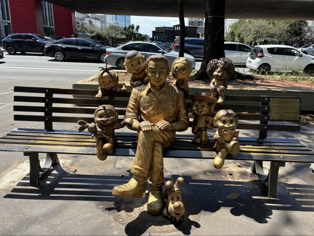 Fotografia diurna, sob sol forte, de uma escultura de bronze dourado em um banco de ripas escuras na calçada de uma avenida movimentada, com árvores e carros ao fundo e um edifício de estrutura vermelha à esquerda. No centro, um homem de bronze está sentado com as pernas cruzadas e as mãos sobre o joelho, sorrindo, de camisa de botões e calça. Ao redor dele, personagens da Turma da Mônica, também em bronze, se agarram ao banco e ao homem: Mônica segurando o Sansão, Cascão, Cebolinha, Magali, Chico Bento de chapéu de palha, Milena e outros. Aos pés da escultura está o cachorrinho Bidu. Uma placa dourada fixada no encosto do banco identifica o monumento.