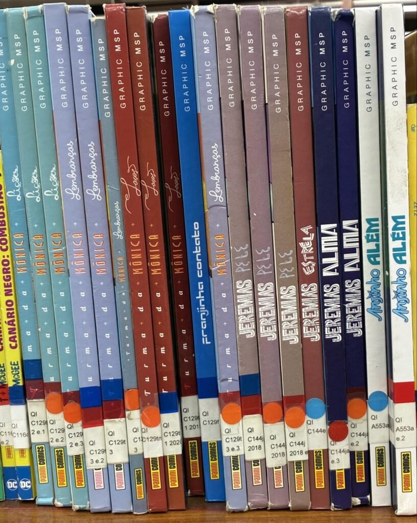 Fotografia de uma prateleira de biblioteca vista de frente, mostrando as lombadas de dezenas de livros enfileirados na vertical, todos da coleção Graphic MSP, da editora Panini Comics. As lombadas variam em cores: azul-turquesa, lilás, cinza-azulado, vermelho escuro, azul vivo, marrom-acinzentado, azul-marinho e branco. Os títulos legíveis incluem “Lições”, “Lembranças” e “Laços”, todos da “Turma da Mônica”, além de “Franjinha: Contato”, “Jeremias: Pele”, “Jeremias: Estrela”, “Jeremias: Alma” e “Anjinho: Além”. Na base de cada lombada há etiquetas brancas de catalogação com códigos como “QI C129t” e “QI C144j”, e adesivos redondos coloridos, em laranja, azul e vermelho. À esquerda aparecem ainda gibis de super-heróis com o logotipo da DC.