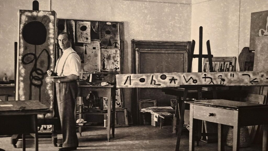 Foto de fotografia em preto e branco do artista surrealista Joan Miró em seu ateliê de arte cercado por telas e esboços.