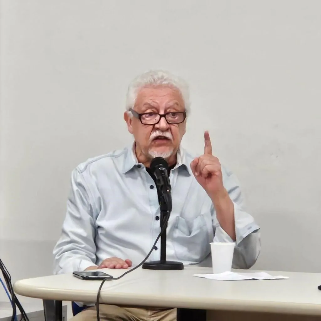 Homem apoia mão direita na mesa e aponta dedo indicador esquerdo para cima.