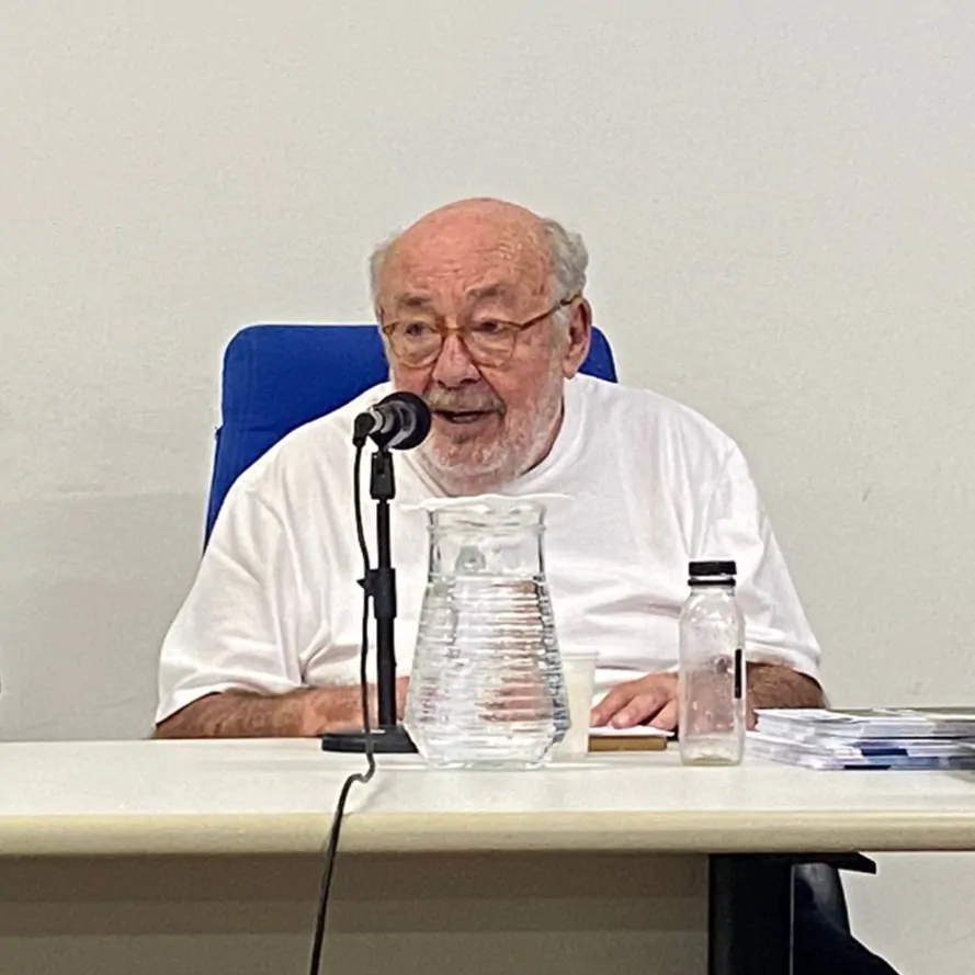 Homem sentado fala ao microfone e segura livro apoiado na mesa.