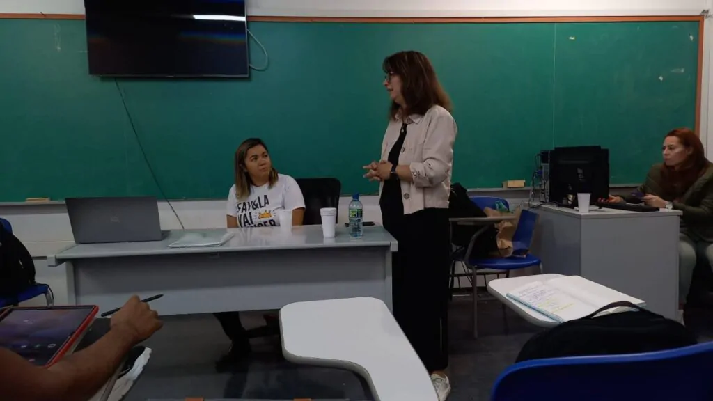 Imagem de uma sala de aula com tr~es mulheres, estando duas sentadas e uma em pé.
