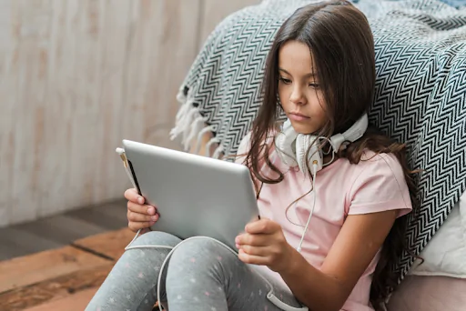 Menina jovem sentada no chão, com fones de ouvido no pescoço, concentrada enquanto usa um tablet, representando o uso precoce e intenso de dispositivos eletrônicos por crianças na era digital.