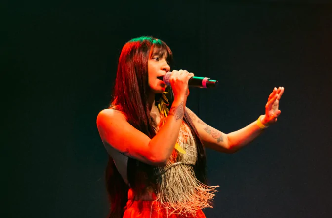A cantora Cayarí está cantando. Voltada para a direita da fotografia, exibe longos cabelos escuros lisos e franja, um top de palha e um shorts vermelho. Ela tem na mão direita um microfone e está iluminada por uma luz vermelha.