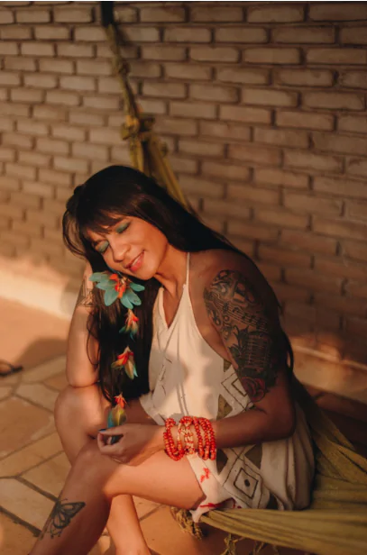A cantora Cayarí, de pele avermelhada e longos cabelos lisos e escuros, está sentada, de olhos fechados, com a cabeça apoiada em sua mão direira, que, por sua vez, está em cima do joelho. Ela está usando sombra azul nos olhos, um brinco de penas, um regata branca e pulseira de bolinhas vermelhas.