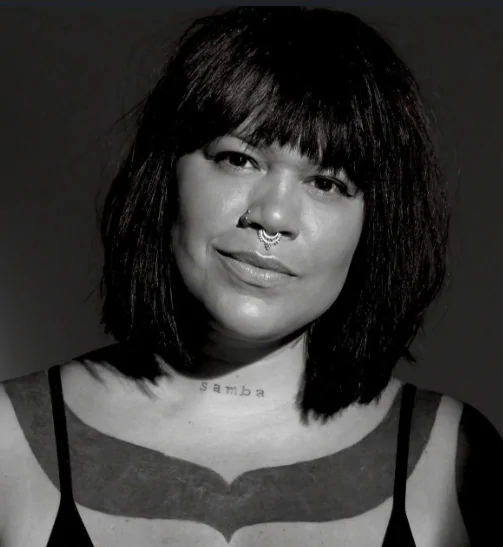 A estilista Dayana Molina, com cabelos escuros lisos na altura do ombro, franja, piercing no septo, regata e tatuagem escrito "samba" no pescoço, encontra-se olhando para frente, com a cabeça levemente inclinada. A foto está em preto e branco.