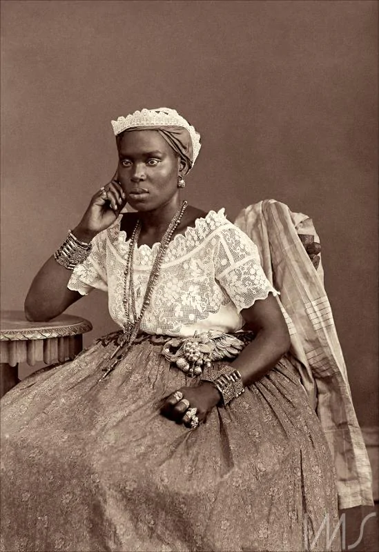 Fotografia de uma mulher negra,com uma longa saia, uma camiseta de renda e um pequeno turbante. Ela está sentada e apoia o cotovelo em uma mesa.