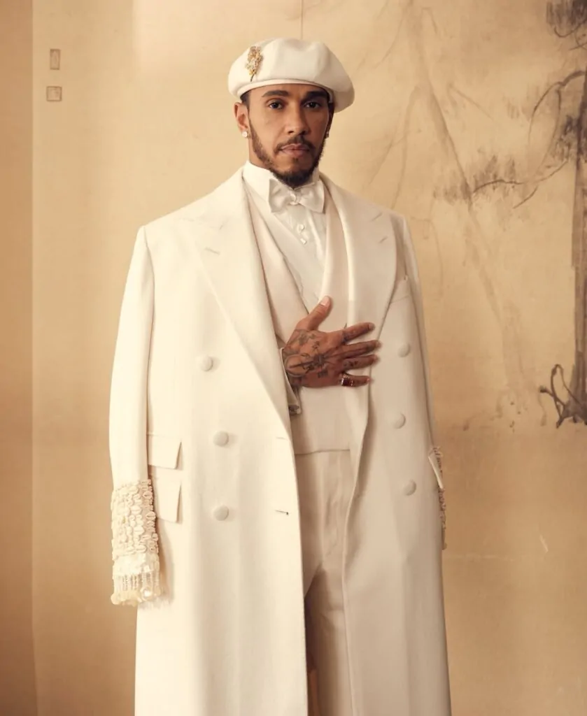 Lewis Hamilton posando com uma roupa inteiramente creme, composta por um calça, camisa, colete, gravat borboleta, um grande paletó aberto e um chapéu de gorro. 