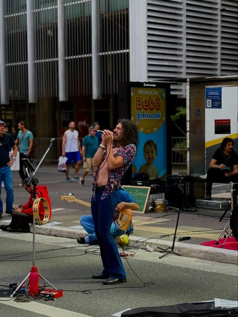 Cantor da banda Glad Zeppelin se apresentando na Paulista. Ele usa uma camisa colorida aberta, uma calça boca de sino azul, sapatos sociais e um cabelo longo solto.
