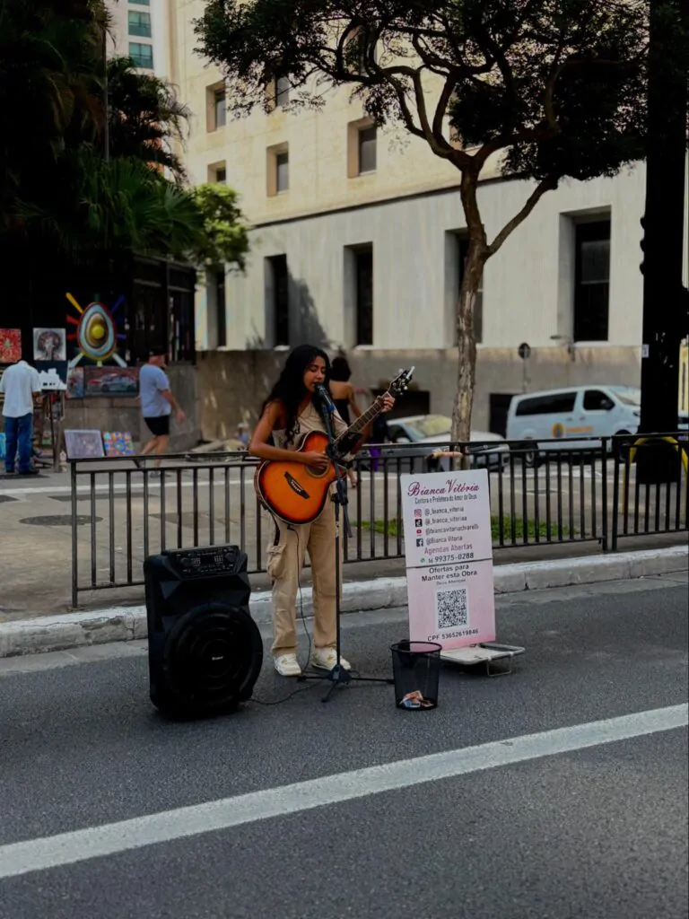 Cantora Bianca Vitória toca violão e canta na Avenida Paulista. Ela usa uma regata bege, uma calça bege de um tom mais escuro e tênis branco.