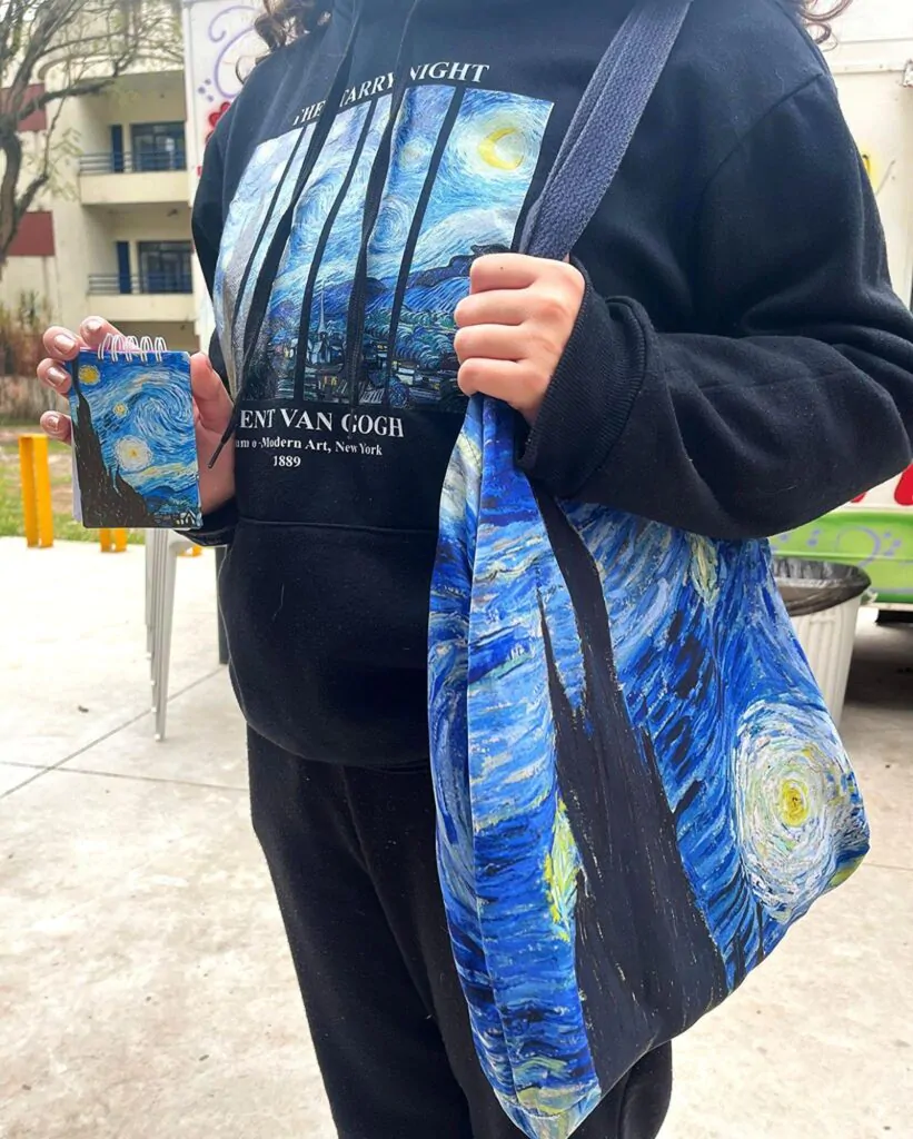 Imagem de uma pessoa vestindo um moletom a estampa da obra "Noite Estrelada", de Vincent Van Gogh. Ela também usa uma bolsa e segura um bloco de notas, ambos com a mesma figura