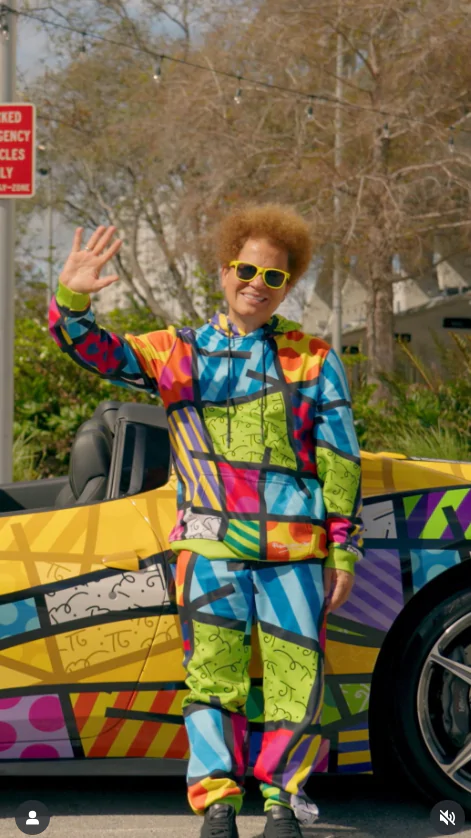 Romero Britto posa na frente de um carro e acena. Suas roupas e o veículos estão estampados no estilo de arte do artista.