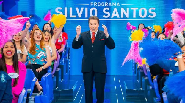 Silvio Santos apresentando o Programa Silvio Santos. Ele usa terno e gravata vermelha e está rodeado de mulheres com pompons.