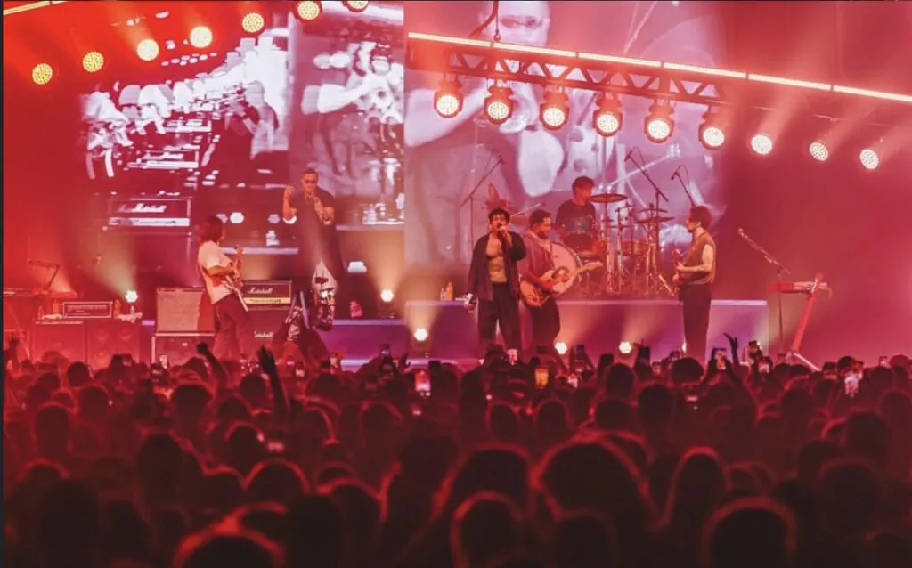 Foto do show da banda Lagum. A imagem é vermelha por conta das luzes, mostra o palco com os artistas e as cabeças da plateia