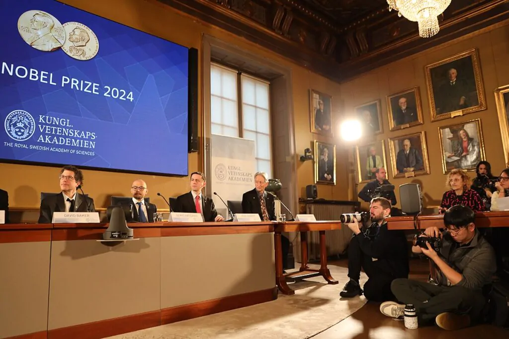 Grupo de pesquisadores reunidos em mesa recebendo prêmio Nobel com fotógrafos ao seu redor.