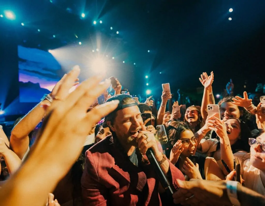 vocalista da banda desce do palco para realizar sua performance próximo à platéia. a imagem é fraterna e passa a atmosfera de carinho dos fãs e humildade do artista. possui luz desfocada ao fundo e muita presença de pessoas