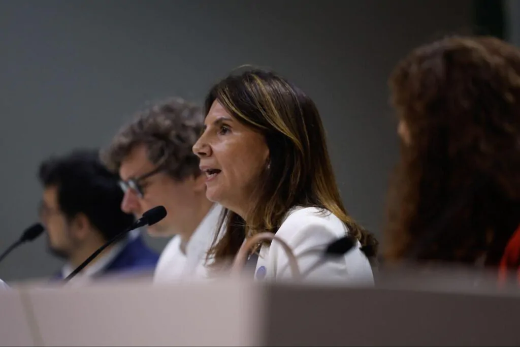 Mulher fala no microfone diante de outras pessoas.