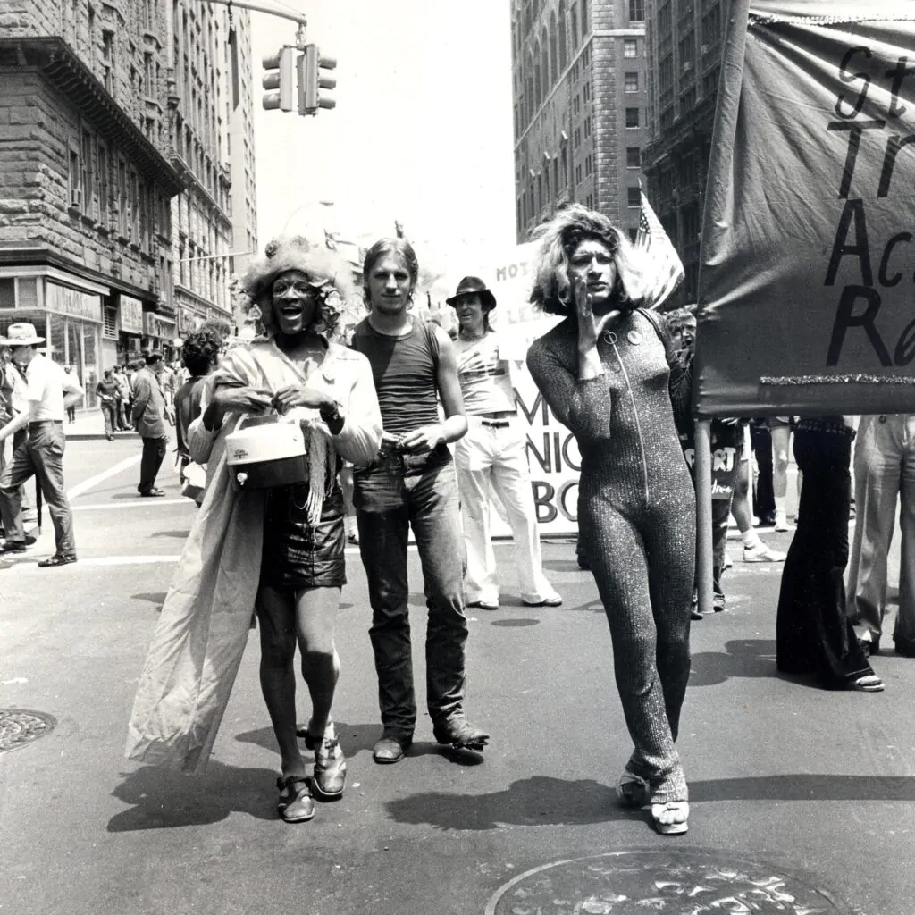 Rivera e Thompson em uma manifestação pacífica em nova York. Ambas aparecem em pé e com roupas belas, Thompson com uma mini saia, camisa e um exuberante chapéu e Rivera com macacão. Thompson possui um grande sorriso na face enquanto Rivera se mostra séria e com a mão na face enquanto segura um lado de um grande cartaz