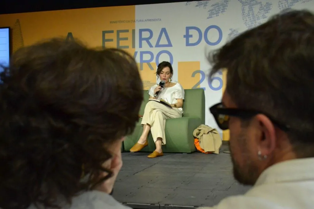 A imagem mostra Maria Brant sentada recitando ao público da Feira do Livro 2026 palavras em um livro