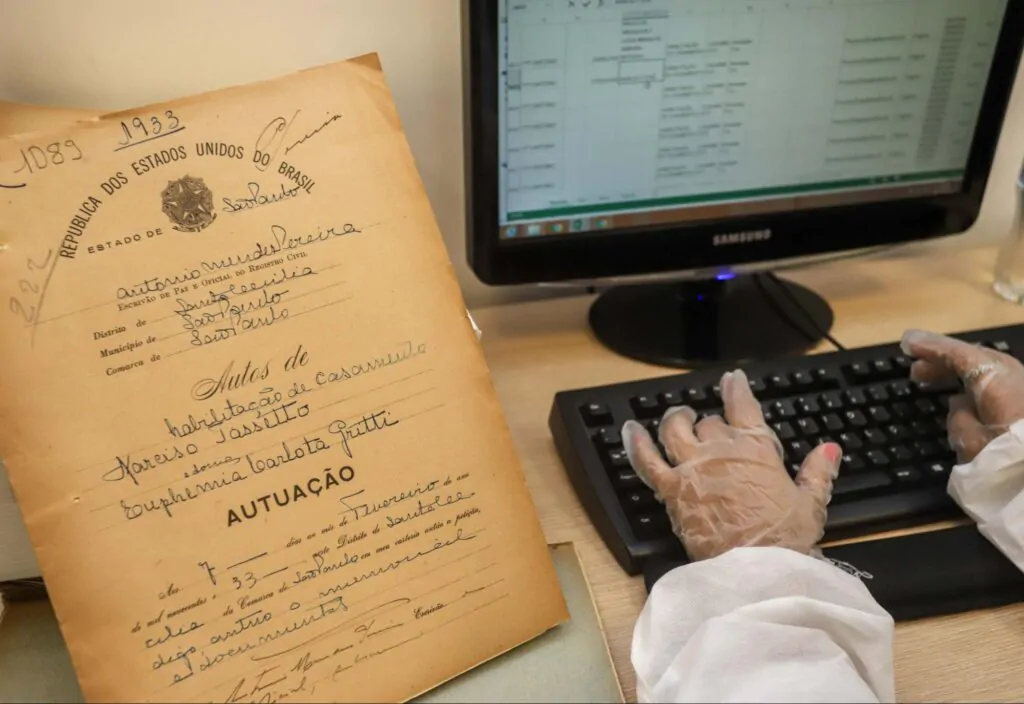 Mãos digitando em um computador informações de um documento antigo.