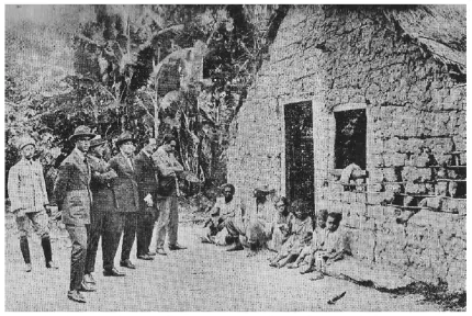 Foto antiga com um grupo de homens olhando construção com crianças e outras pessoas sentadas na frente.