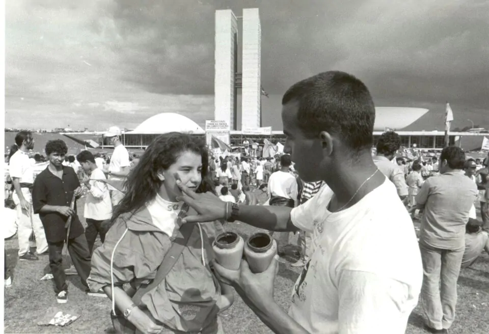 Homem pardo de camisa branca pintando o rosto de uma mulher branca e de cabelos longos