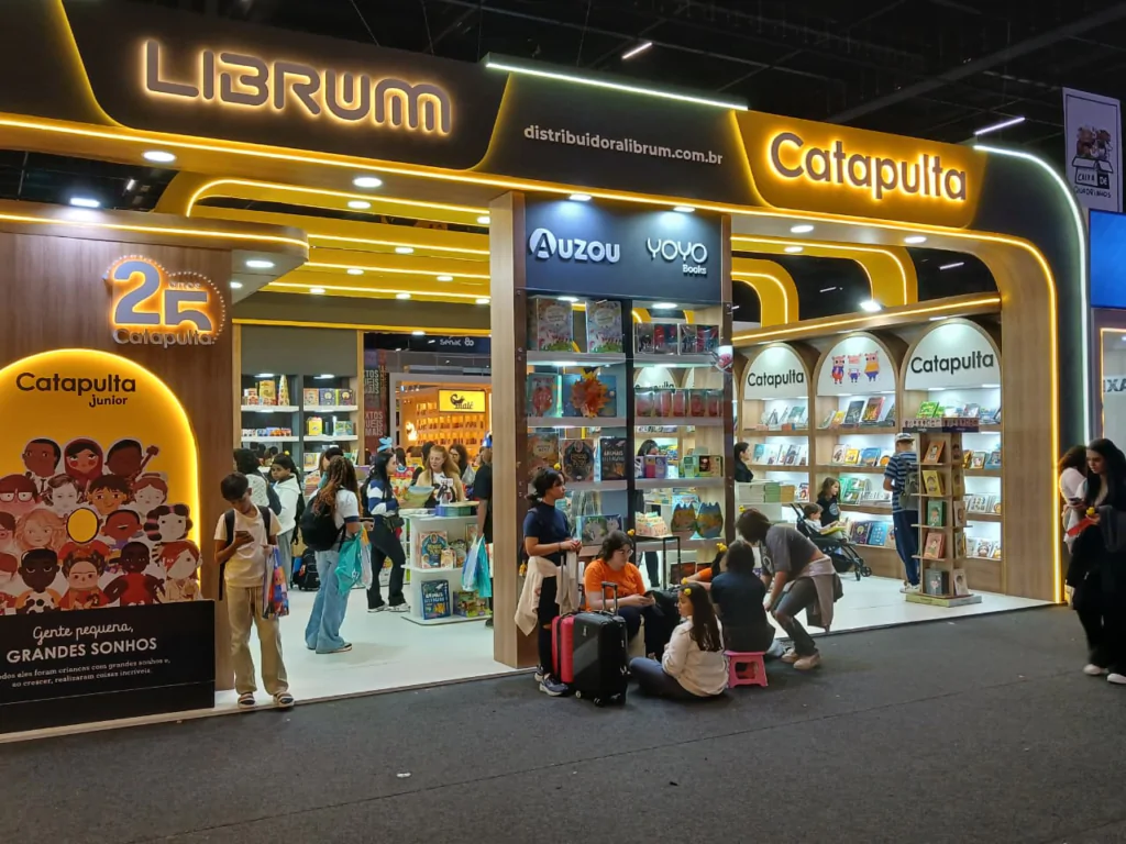 Fotografia em plano geral e iluminação artificial de um estande moderno e bem iluminado da editora Librum e Catapulta em uma feira de livros. O estande possui estrutura de madeira com detalhes curvados e iluminação em fitas de LED amareladas. Na fachada superior, destacam-se os letreiros luminosos em amarelo com os nomes "LIBRUM" e "Catapulta". À esquerda, um painel ilustrado exibe o selo "25 anos Catapulta" acima da frase "Gente pequena, GRANDES SONHOS". No centro e ao fundo, estantes repletas de livros infantis atraem visitantes de diversas idades que transitam pelo local. Em primeiro plano, no corredor do pavilhão, um pequeno grupo de pessoas está sentado no chão descansando ao lado de malas de viagem, enquanto outros clientes observam as prateleiras e caminham pelo espaço.