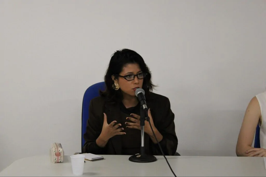 Mulher negra de óculos, blazer marrom falando ao microfone