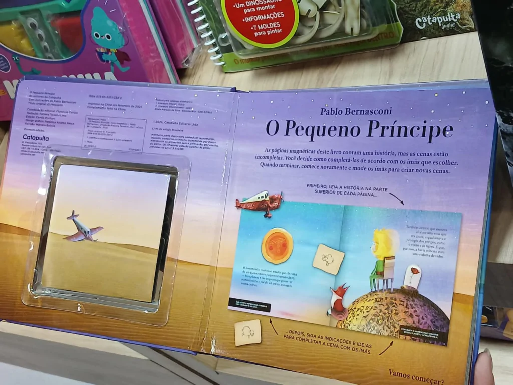 Fotografia em plano médio tirada de cima, mostrando um livro infantil ilustrado aberto sobre uma mesa clara, intitulado "O Pequeno Príncipe", escrito por Pablo Bernasconi. A página da esquerda possui no topo textos com informações de créditos editoriais e, na parte central, um quadro magnético retangular que simula uma paisagem desértica sob céu claro, no qual há um pequeno ímã em formato de avião posicionado do lado esquerdo. A página da direita é de tom azul-claro estrelado e traz o nome do autor e o título em destaque. Abaixo, há instruções de como brincar com os ímãs magnéticos do livro, ilustradas com exemplos de peças como um avião vermelho, a figura do Pequeno Príncipe em seu planeta e a raposa sobre o solo arenoso. Ao fundo, na parte superior da imagem, veem-se outros produtos e cadernos infantis coloridos parcialmente expostos.
