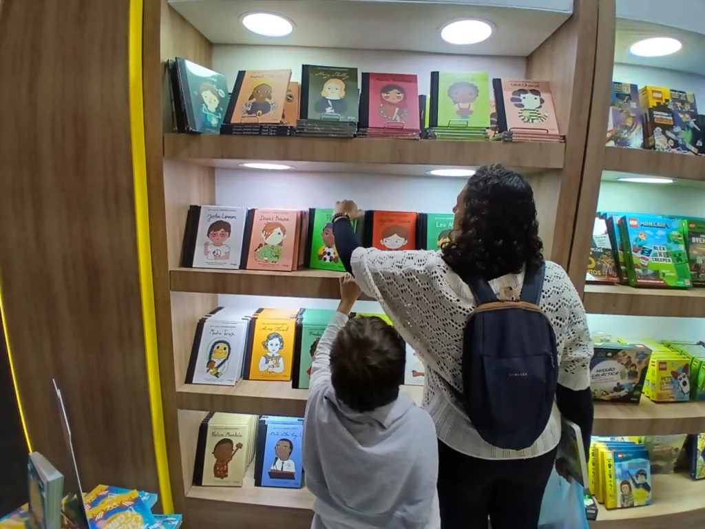 Fotografia em plano médio tirada do interior de uma livraria, mostrando uma mulher e uma criança de costas enquanto observam livros em uma estante de madeira iluminada. A mulher, situada à direita, usa uma blusa de tricô furadinha na cor creme sobre uma peça de mangas compridas escura e carrega uma mochila azul-marinho nas costas. Ela está com o braço direito estendido alcançando um livro na segunda prateleira de cima para baixo. A criança, posicionada à esquerda e um pouco à frente da mulher, veste um casaco de moletom cinza claro e estende o braço esquerdo em direção à terceira prateleira. A estante possui quatro prateleiras principais repletas de livros infantis ilustrados, dispostos de frente para exibição das capas com fundos coloridos e retratos de personagens. Do lado direito e em uma mesa no canto inferior esquerdo, há outros exemplares e caixas de jogos empilhados sob a iluminação de lâmpadas embutidas no teto do móvel.