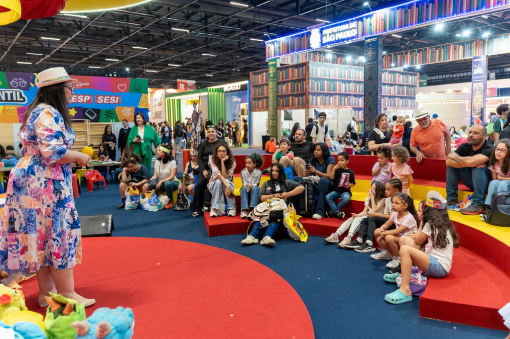 Fotografia em plano geral e iluminação ampla de um espaço infantil em uma feira de livros. À esquerda, em primeiro plano, uma mulher de chapéu e vestido longo florido em tons de azul, rosa e branco está em pé sobre um tapete circular vermelho, voltada para o público. Diante dela, dezenas de crianças e adultos estão acomodados em arquibancadas coloridas em formato semicircular, nas cores amarela e vermelha, dispostas sobre um piso azul-escuro. A plateia assiste atenta à atividade, com várias crianças sentadas nos degraus mais baixos. Ao fundo, sob a iluminação do pavilhão de eventos, veem-se estandes decorados, incluindo a estrutura da "PREFEITURA DE SÃO PAULO" ornamentada com estampas de prateleiras de livros e o espaço "ESP SESI".